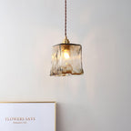 Square Clérachic glass pendant lit against a pale wall, with a framed card on a ledge and a brown twisted cord above it.