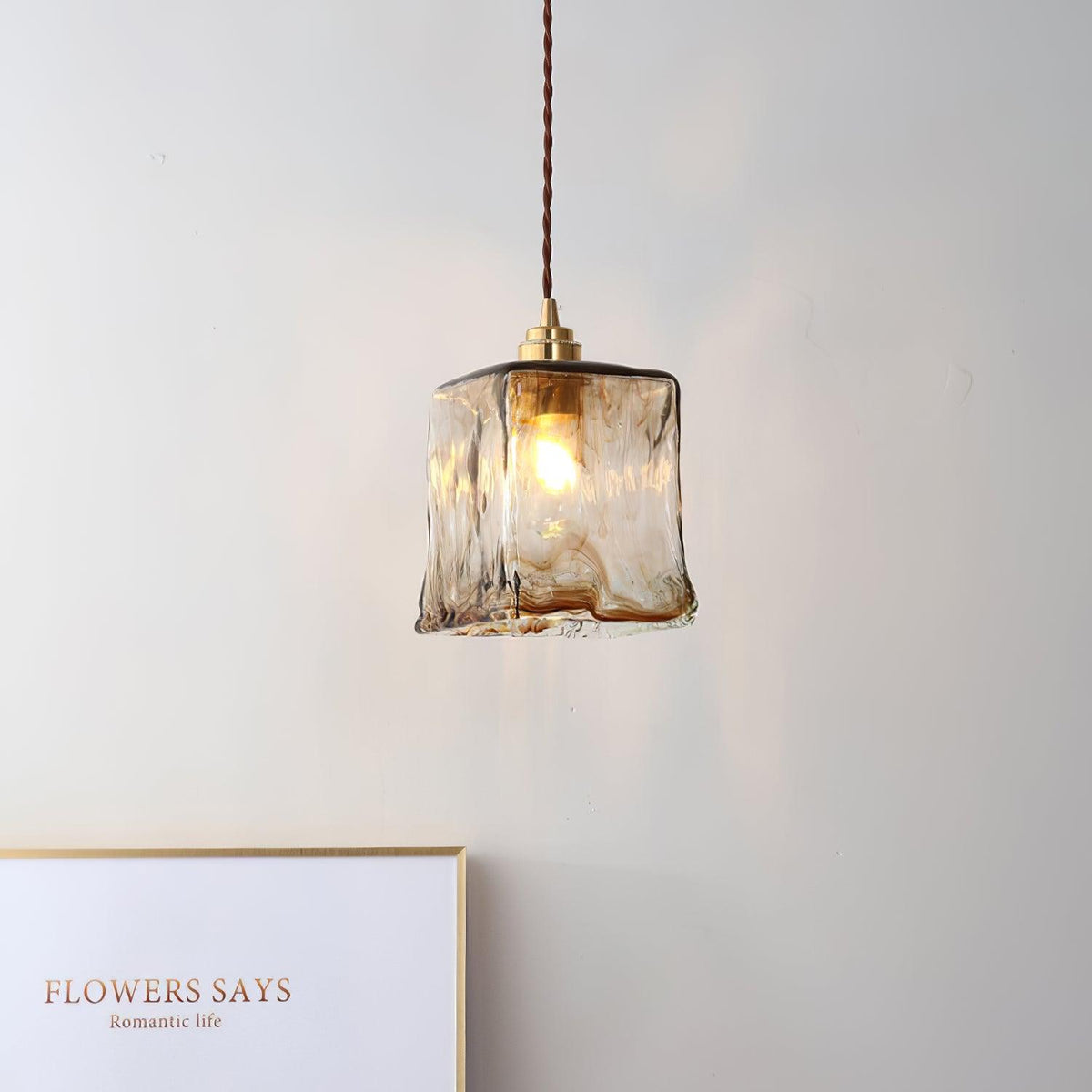 Square Clérachic glass pendant lit against a pale wall, with a framed card on a ledge and a brown twisted cord above it.