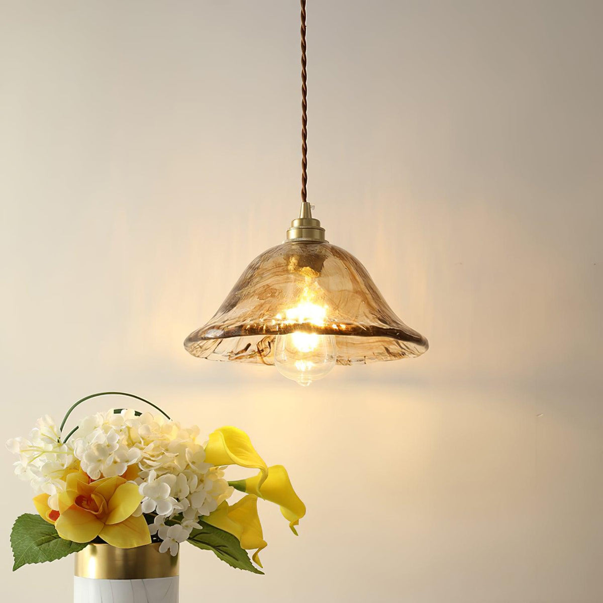 Clérachic bell-shaped brown glass pendant glowing beside a bunch of yellow and white flowers against a cream wall.