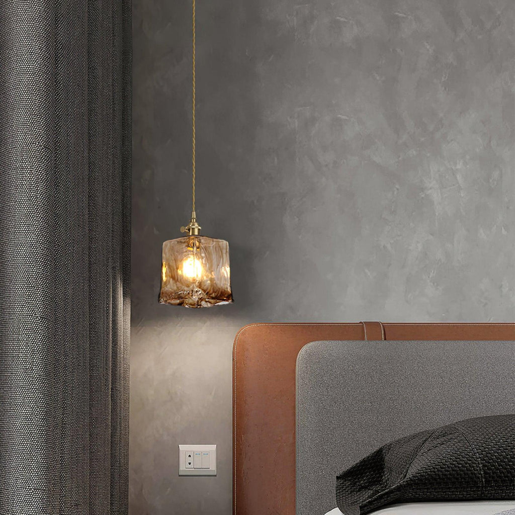 A small Clérachic cube pendant in swirled brown glass lit over a bed, with a rust leather headboard and a dark grey curtain.