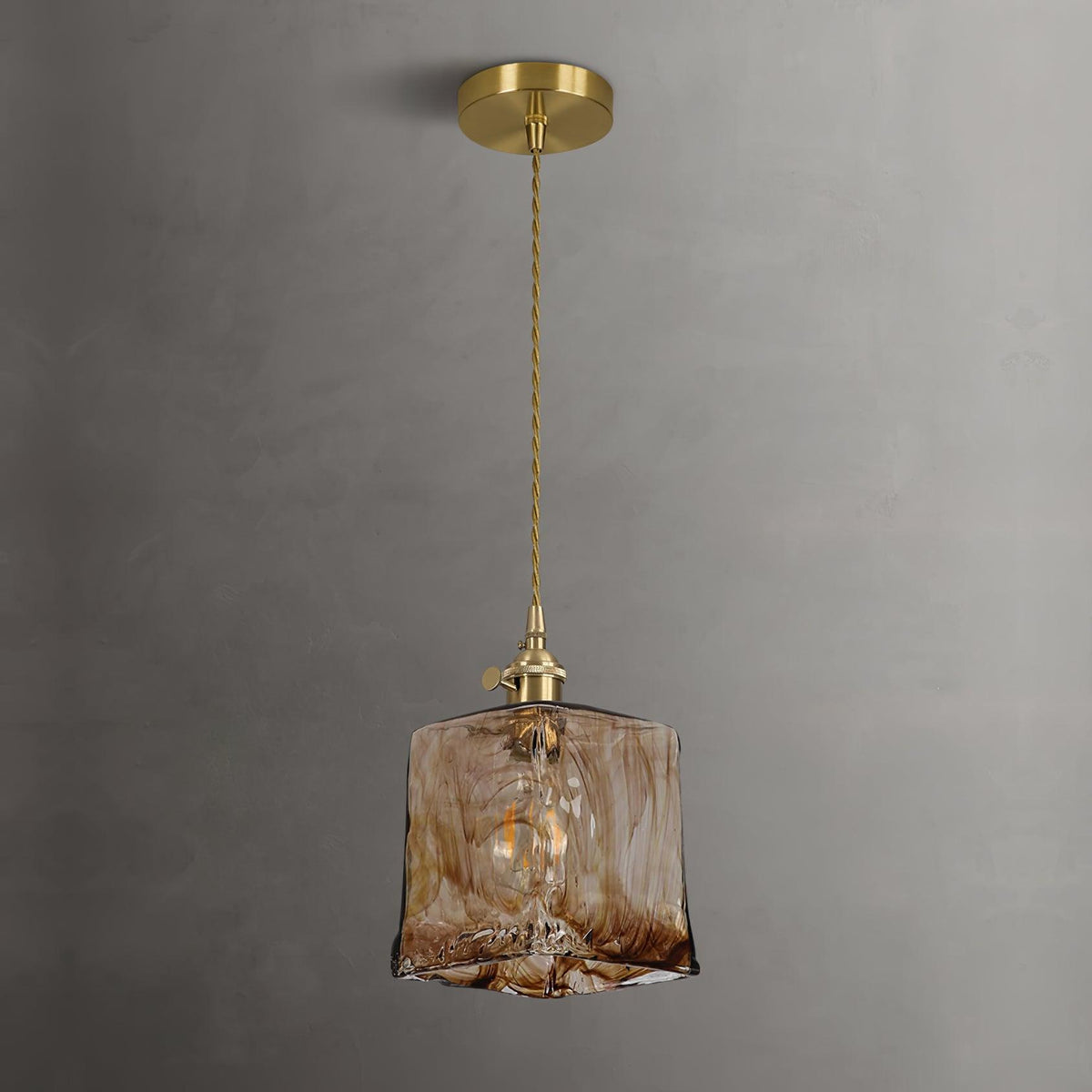 Clérachic cube pendant in swirled brown glass on a brass ceiling plate and twisted cord, hung against a grey plaster wall.