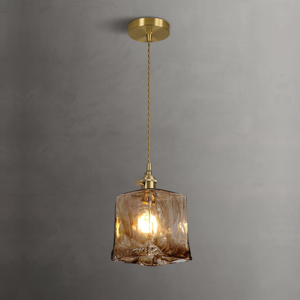 Lit Clérachic cube pendant on a brass ceiling plate against a grey wall, with warm light through the swirled glass.