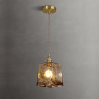 Lit Clérachic cube pendant on a brass ceiling plate against a grey wall, with warm light through the swirled glass.