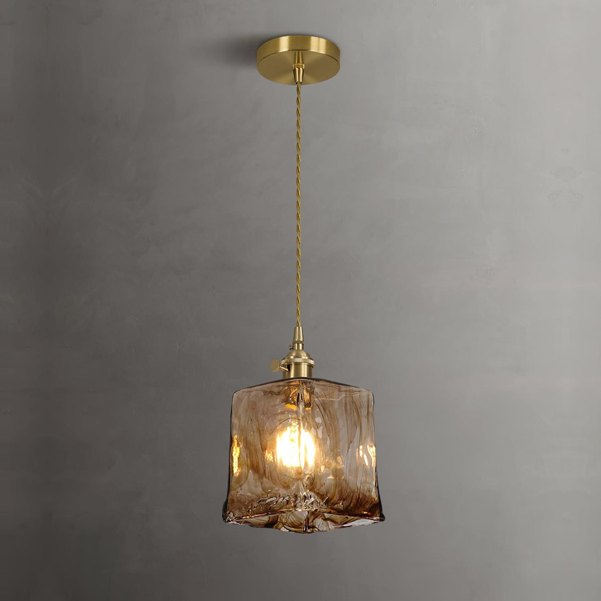 Lit Clérachic cube pendant on a brass ceiling plate against a grey wall, with warm light through the swirled glass.