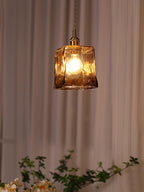 Clérachic cube pendant lit in a dusty-pink curtained room, with the amber glass glowing above a few green leaves.
