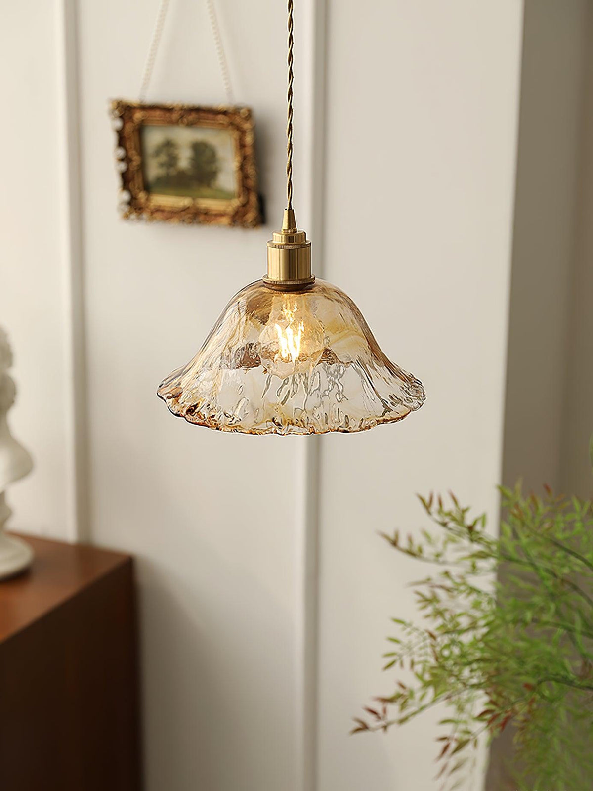 A Clérachic bell-shaped pendant in pale, rippled amber glass hung on a white wall beside a small gilt-framed picture.