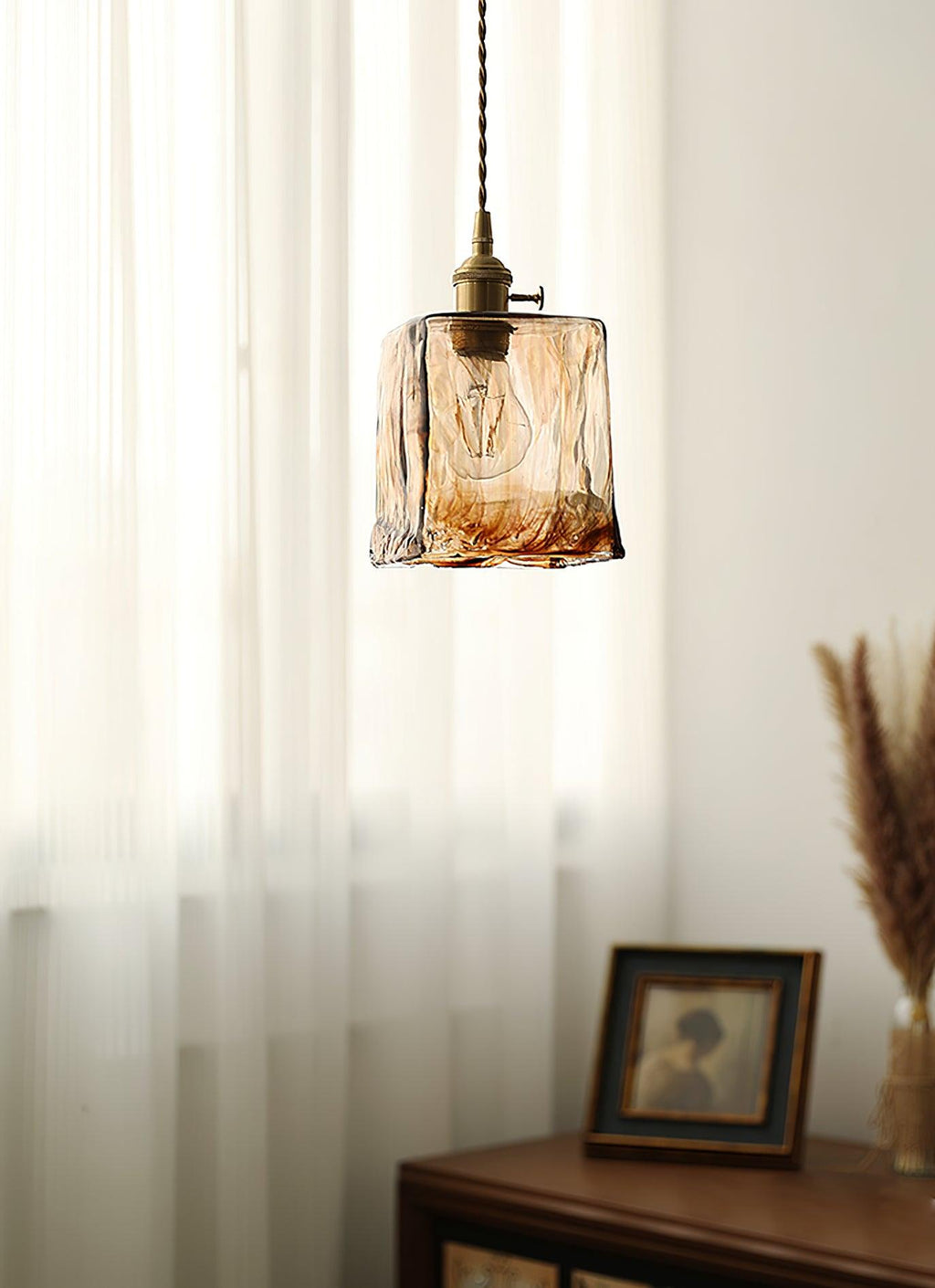 Clérachic cube pendant in rippled brown glass in front of sheer curtains, with a framed portrait and a dried grass stem to the right.