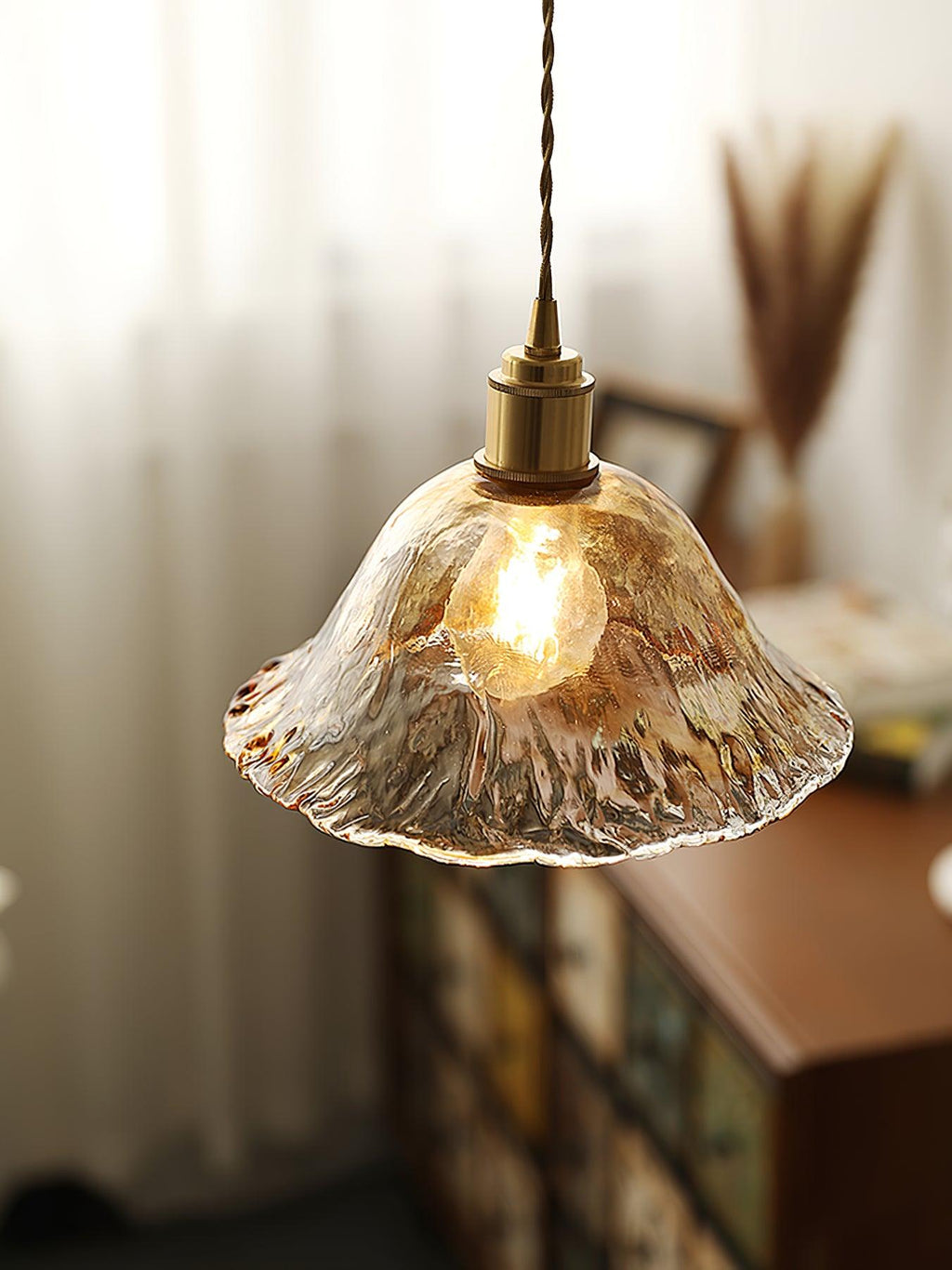 Close view of this Clérachic bell pendant in textured clear-amber glass, with a brass socket and a warm filament bulb, by a window.