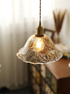 Close view of this Clérachic bell pendant in textured clear-amber glass, with a brass socket and a warm filament bulb, by a window.