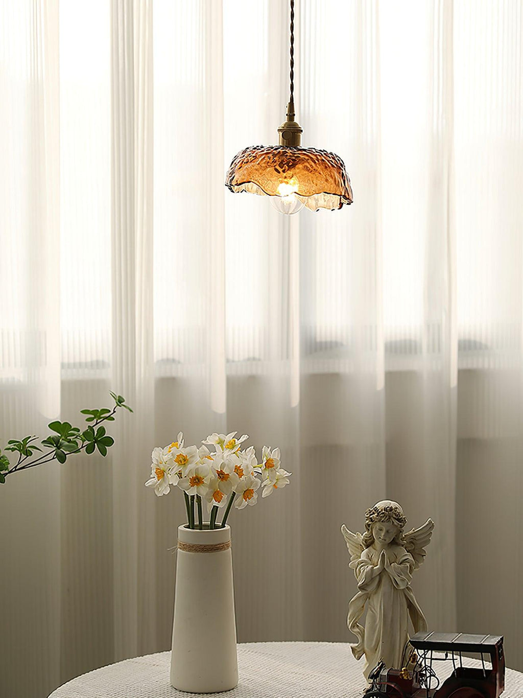 Small Clérachic scalloped-dome pendant in amber glass in front of sheer curtains, above a vase of narcissi and a small angel figure.