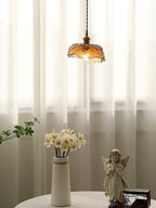 Small Clérachic scalloped-dome pendant in amber glass in front of sheer curtains, above a vase of narcissi and a small angel figure.