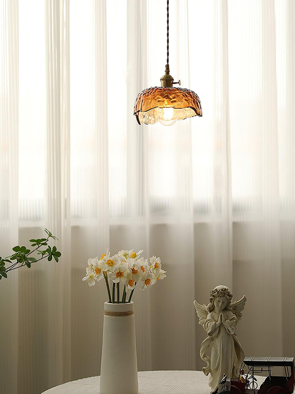 Clérachic scalloped-dome glass pendant in front of sheer curtains, with a vase of narcissi and an angel figure on a round table below.