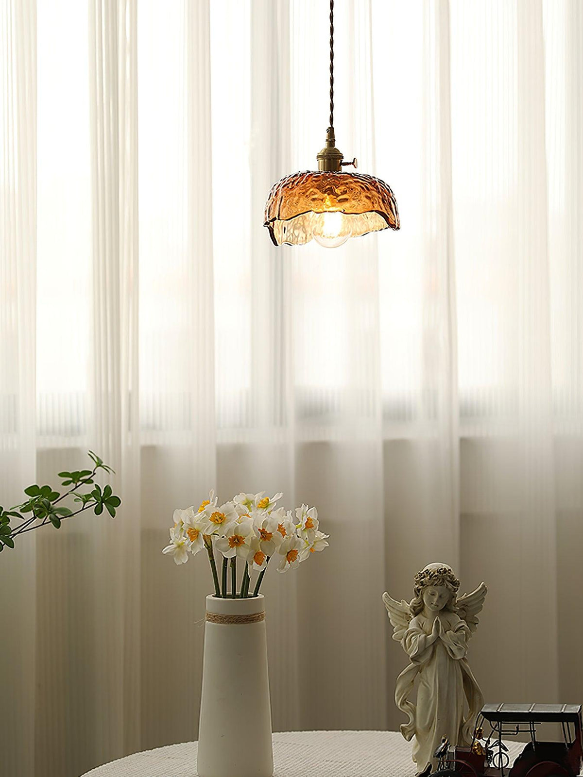 Clérachic scalloped-dome glass pendant in front of sheer curtains, with a vase of narcissi and an angel figure on a round table below.