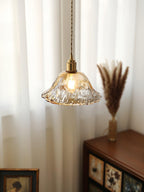 A Clérachic bell pendant in pale rippled glass, lit in front of a white curtain with a small wooden cabinet and dried grass.