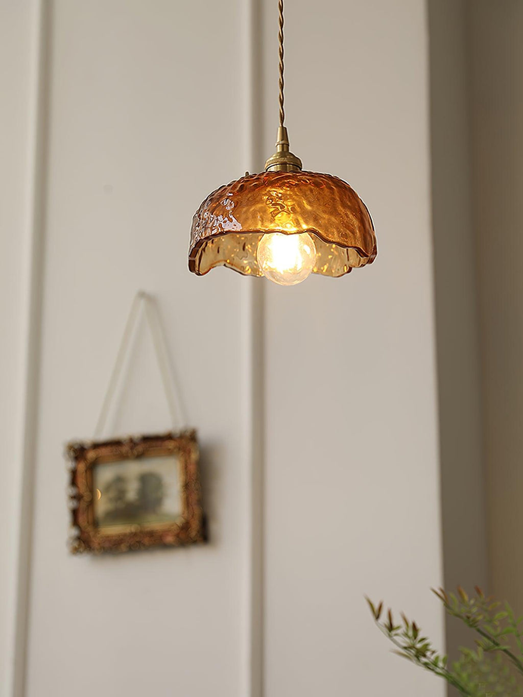 This Clérachic dome-shaped pendant in hammered amber glass, with a brass socket and switch, beside a framed picture on a white wall.