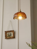 This Clérachic dome-shaped pendant in hammered amber glass, with a brass socket and switch, beside a framed picture on a white wall.