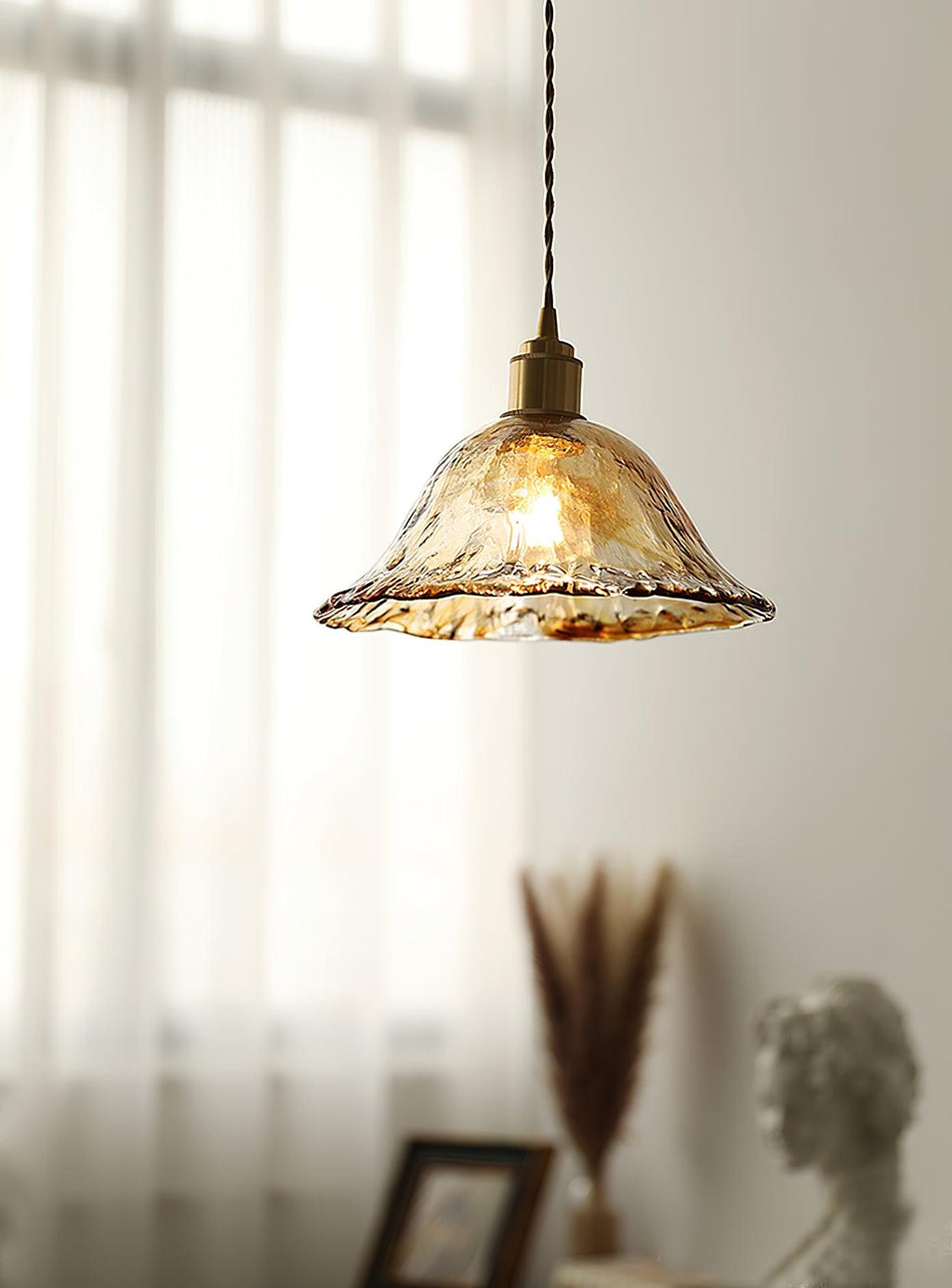 Clérachic bell pendant in clear, rippled glass with a gold-brown rim, lit against sheer curtains above a dried grass arrangement.