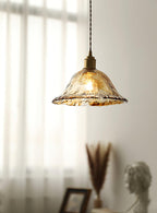 Clérachic bell pendant in clear, rippled glass with a gold-brown rim, lit against sheer curtains above a dried grass arrangement.