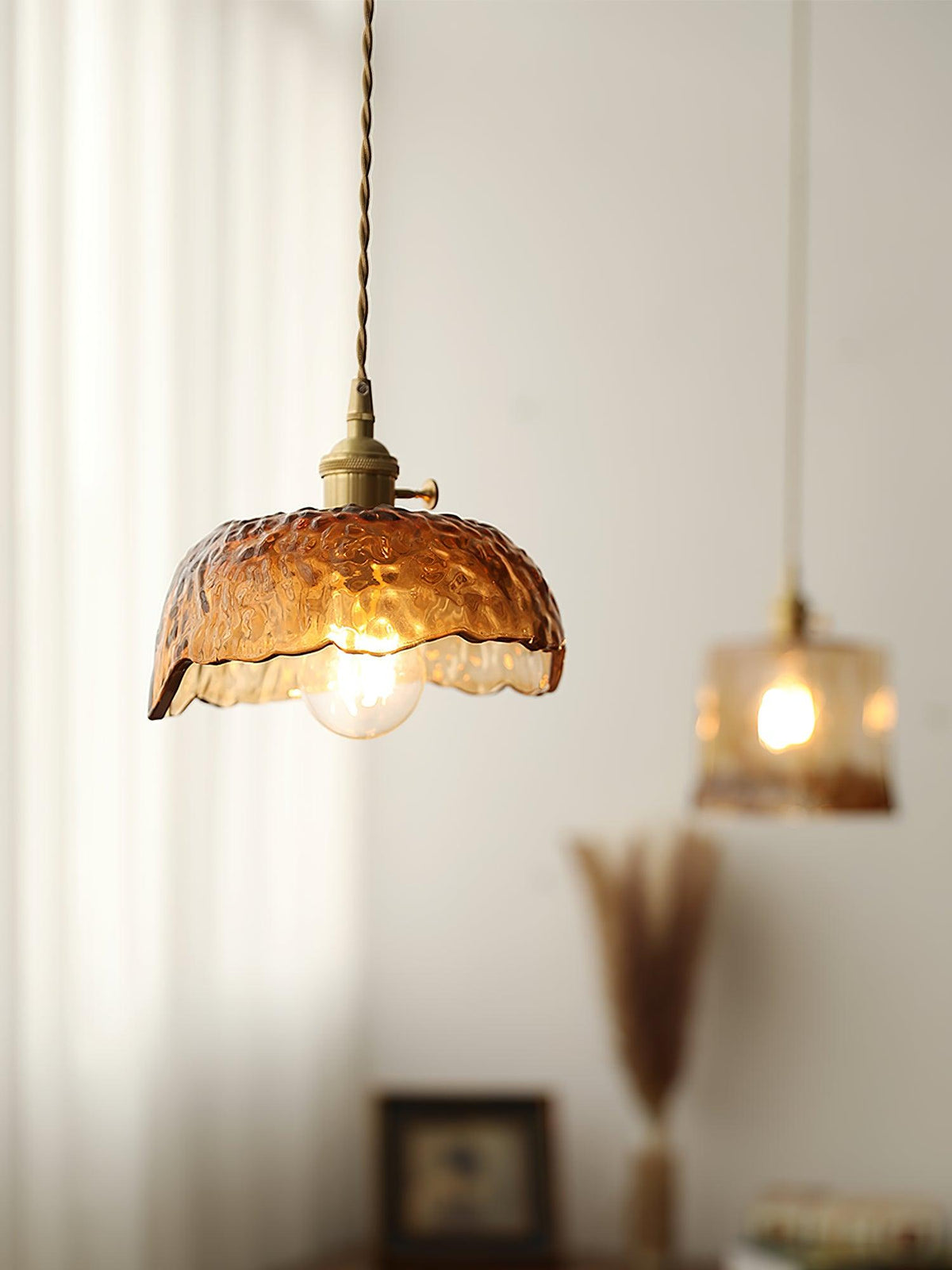 A Clérachic hammered-glass dome pendant in the foreground with a second small glass pendant glowing behind it, in a curtained room.