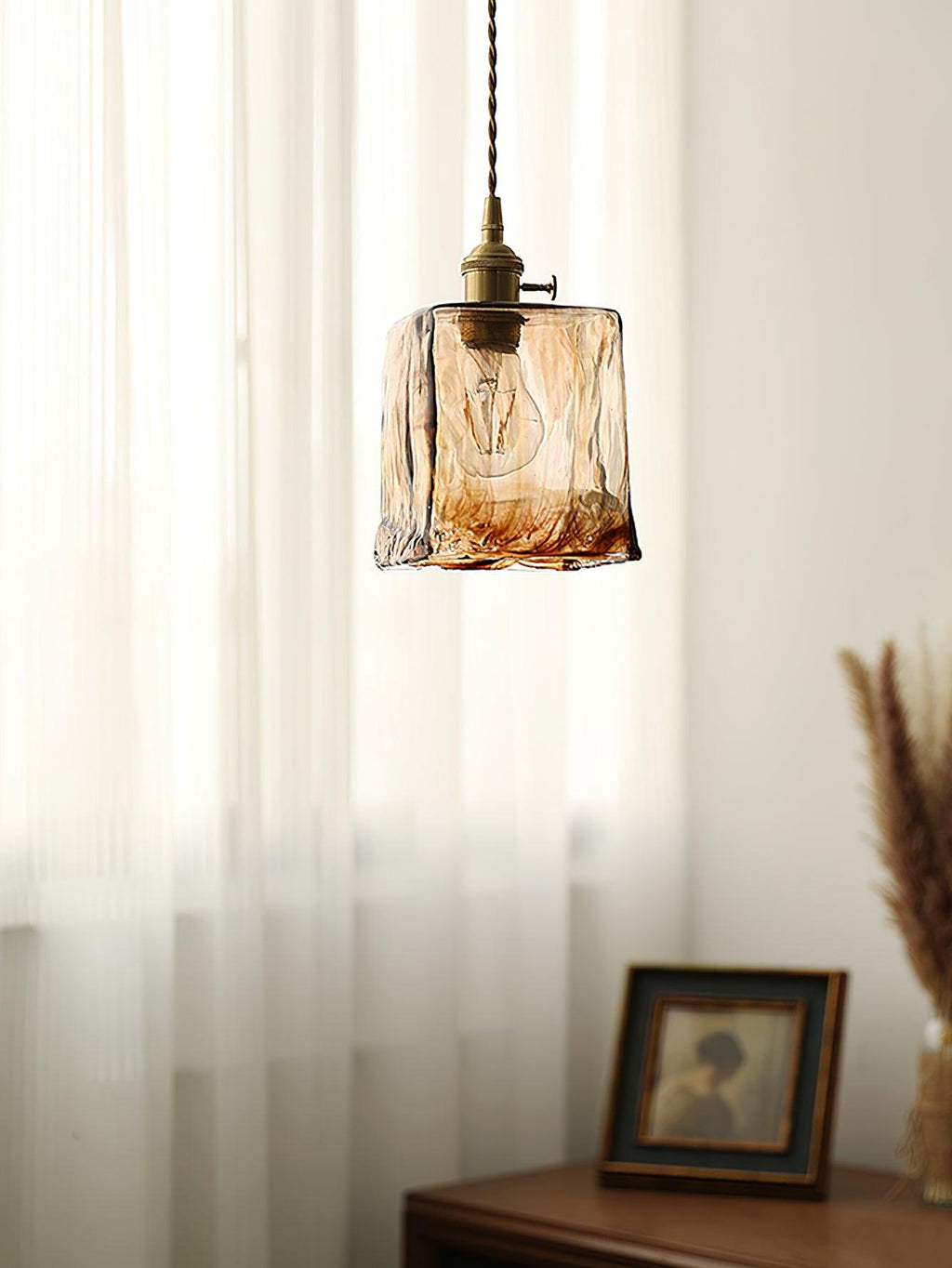 Clérachic cube pendant in swirled brown glass in front of sheer curtains, with a framed portrait on a wooden cabinet below.