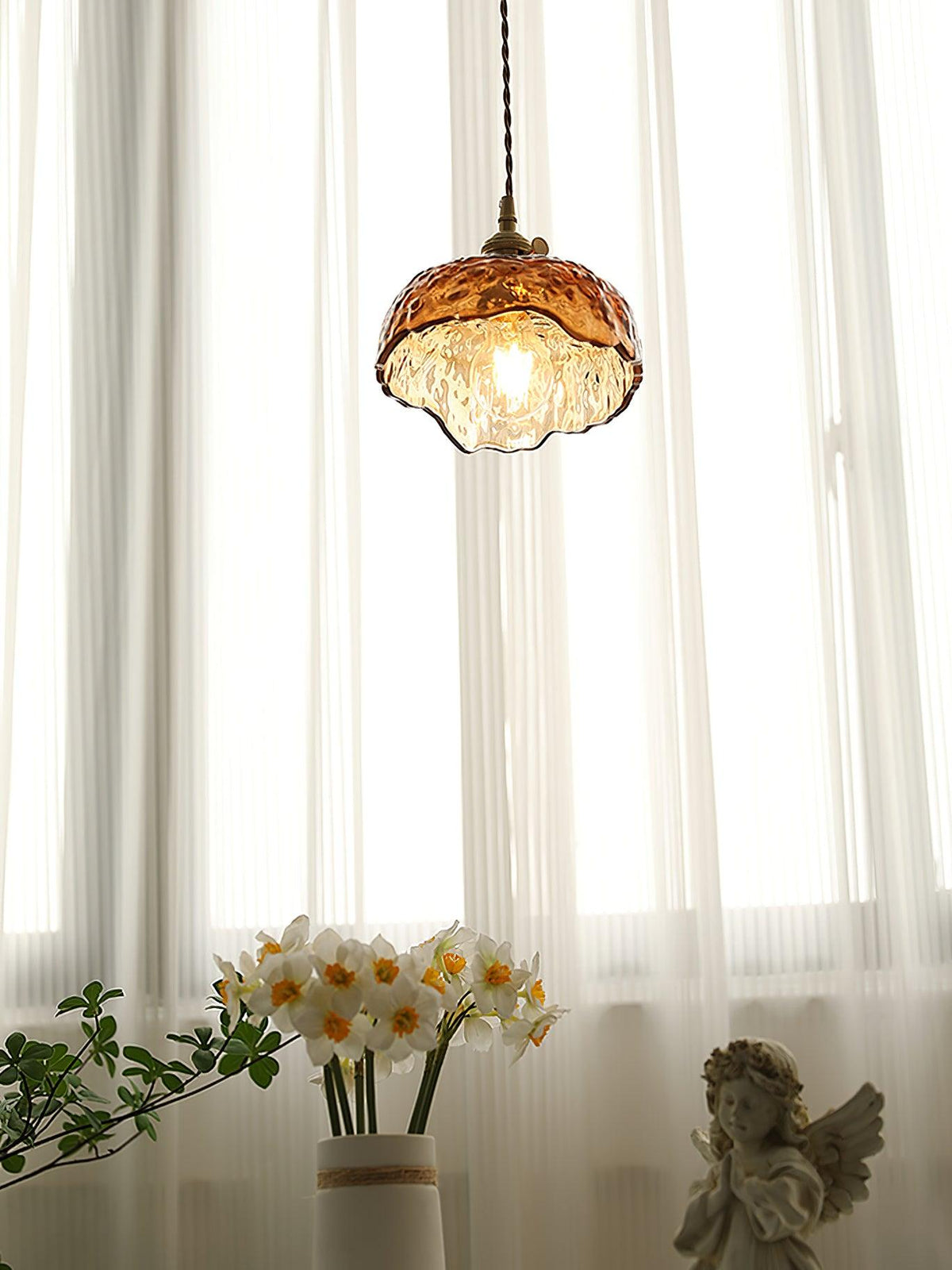 Small Clérachic dome pendant in hammered amber glass, lit above a vase of white narcissi and a figure, in front of tall curtains.