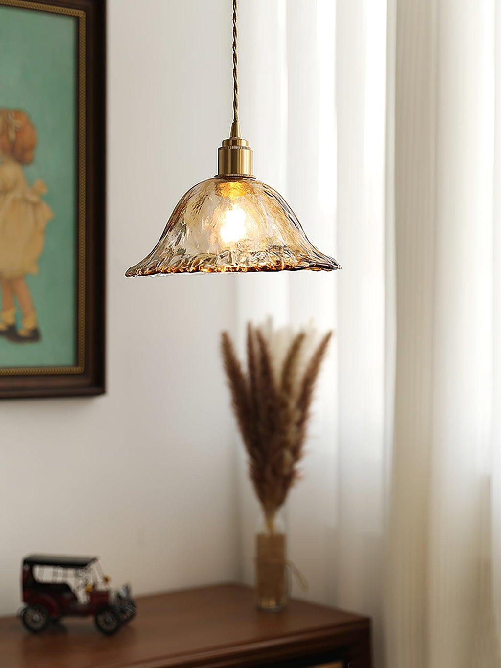 A Clérachic bell pendant in rippled amber glass, hung in front of a framed painting and a stem of dried grass on a wooden cabinet.