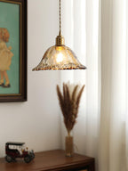 A Clérachic bell pendant in rippled amber glass, hung in front of a framed painting and a stem of dried grass on a wooden cabinet.