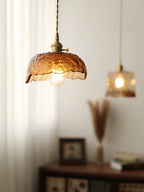 Clérachic dome pendant in hammered amber glass with a cube pendant glowing behind it, beside a dried grass vase and a small cabinet.
