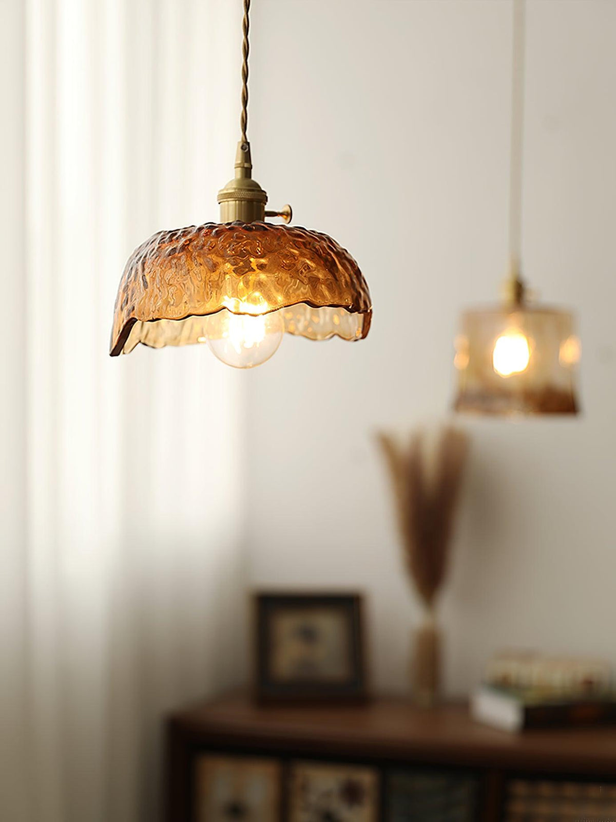 Clérachic dome pendant in hammered amber glass with a cube pendant glowing behind it, beside a dried grass vase and a small cabinet.