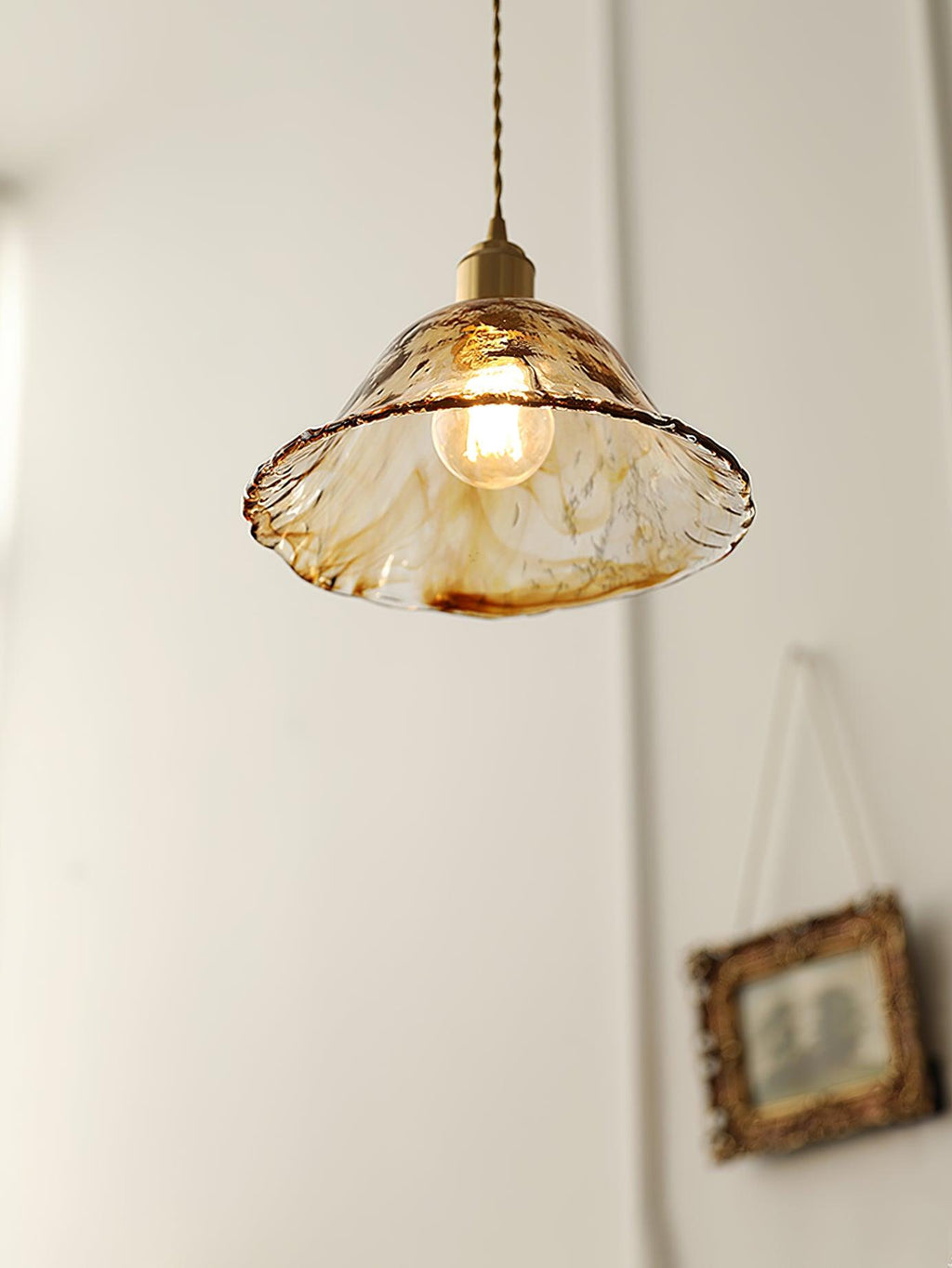 Clérachic bell pendant in brown glass with a frilled rim, lit against a white wall beside a hanging gilt picture frame.
