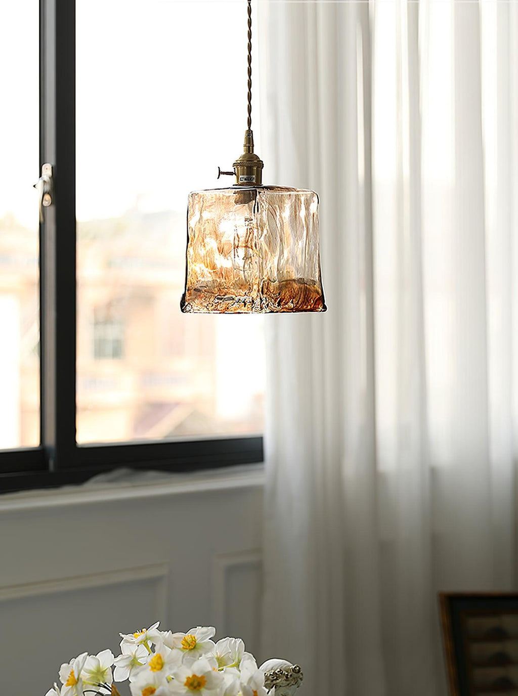 A Clérachic cube pendant in rippled brown glass hung by a window with sheer curtains and a vase of white narcissi below.