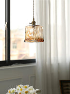 A Clérachic cube pendant in rippled brown glass hung by a window with sheer curtains and a vase of white narcissi below.