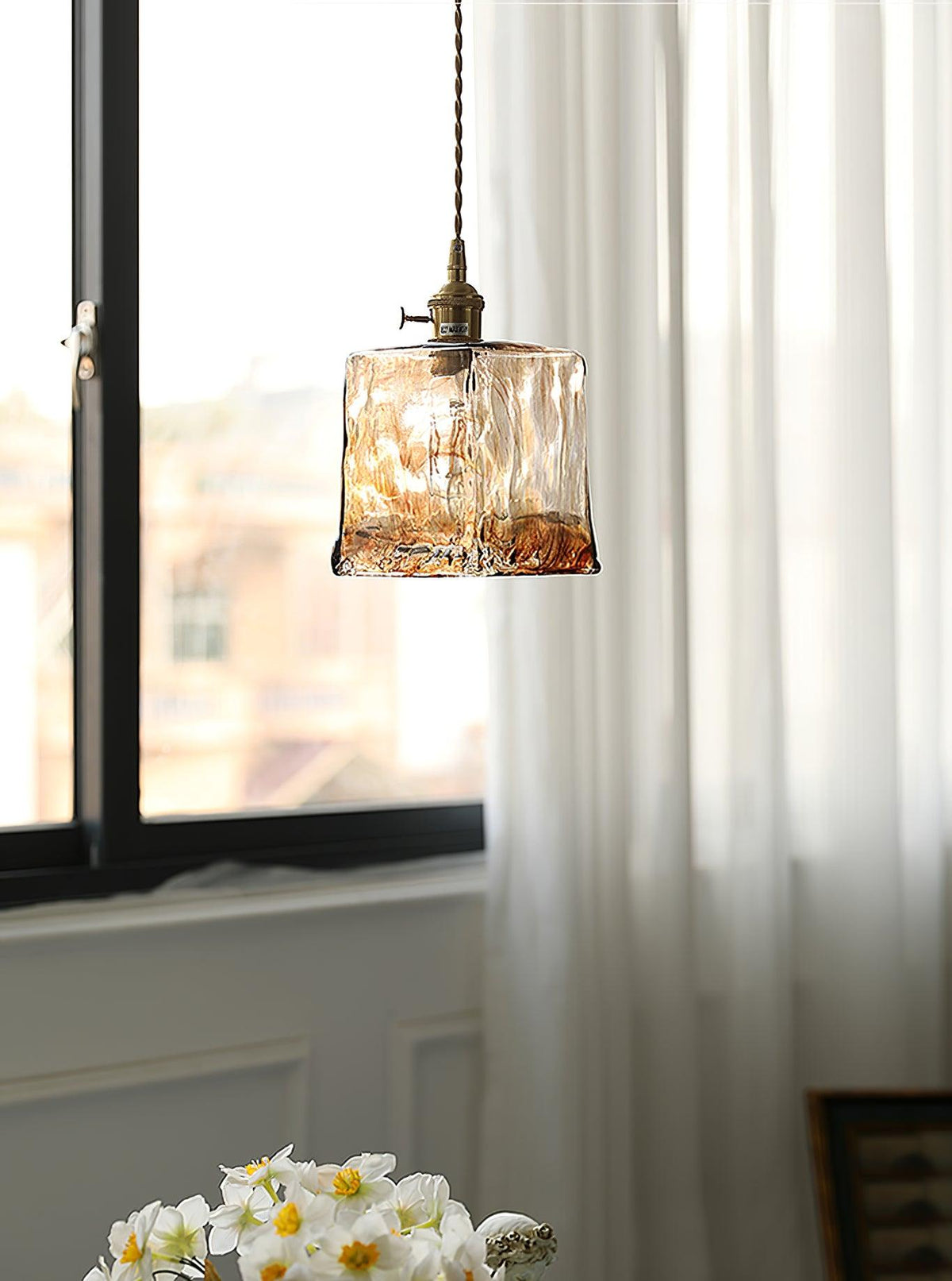 A Clérachic cube pendant in rippled brown glass hung by a window with sheer curtains and a vase of white narcissi below.