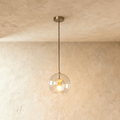 Clérachic pendant light with a clear ribbed glass globe and brass fitting, hung on a black cord from a plaster ceiling.