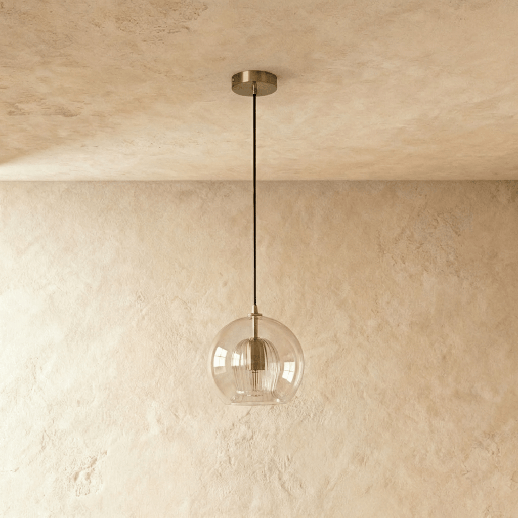 Clérachic pendant light, clear glass sphere over a fluted inner shade, brass ceiling rose against a textured wall.