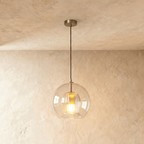 Clérachic pendant light lit warm, its clear glass globe and fluted inner shade glowing against a plaster wall.
