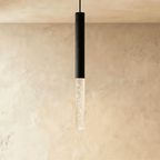 Clérachic pendant with a black cylindrical body and a longer bubble acrylic shade, glowing softly on a limewash wall.