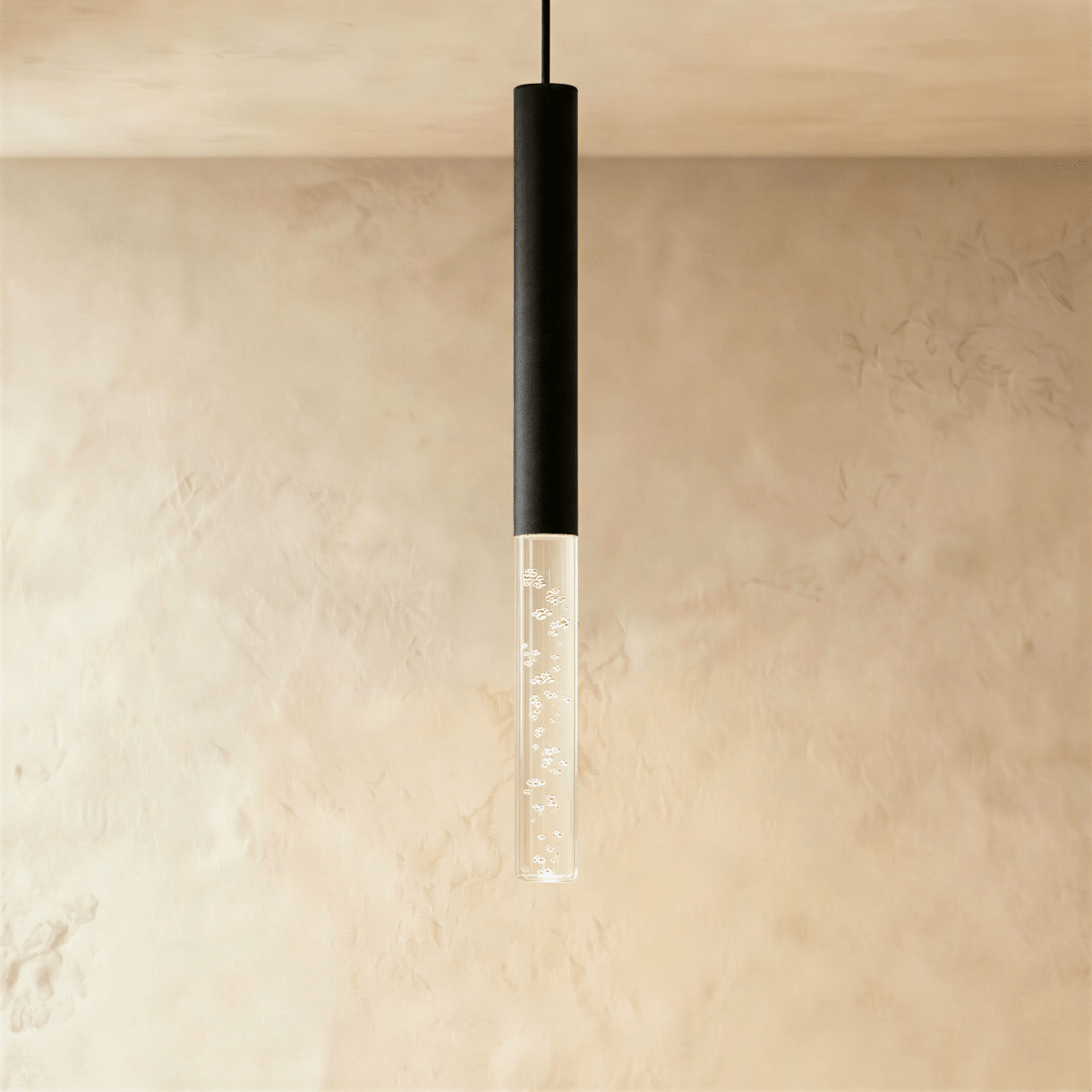 Clérachic pendant with a black cylindrical body and a longer bubble acrylic shade, glowing softly on a limewash wall.