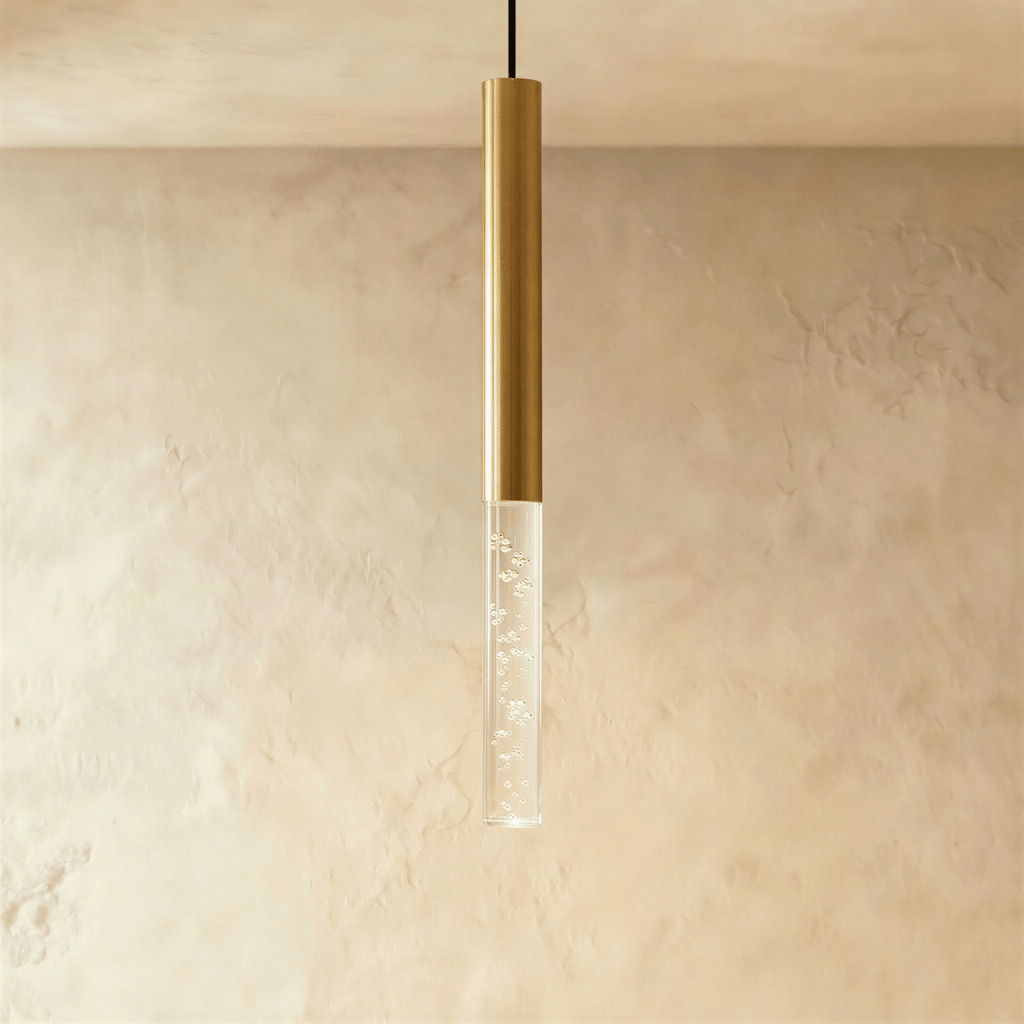 Clérachic gold cylinder pendant with a longer clear acrylic shade holding tiny bubbles, hung before a plaster wall.