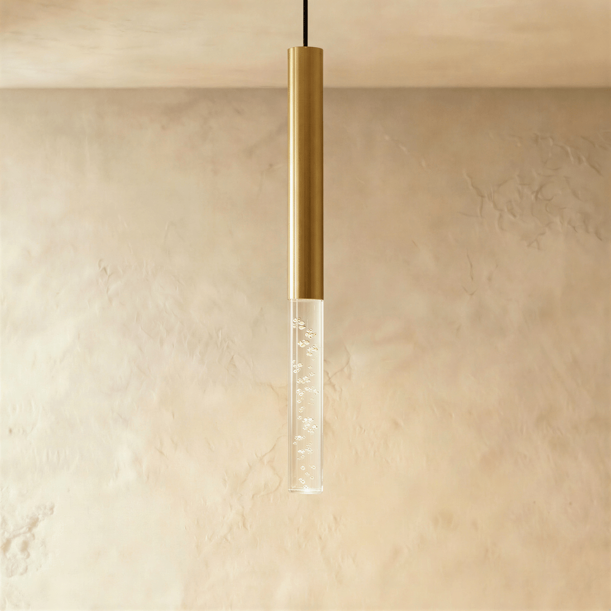 Clérachic gold cylinder pendant with a longer clear acrylic shade holding tiny bubbles, hung before a plaster wall.
