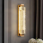 Lit bubble-crystal wall light from Clérachic on a grey panelled wall beside dark curtains, with white flowers below.