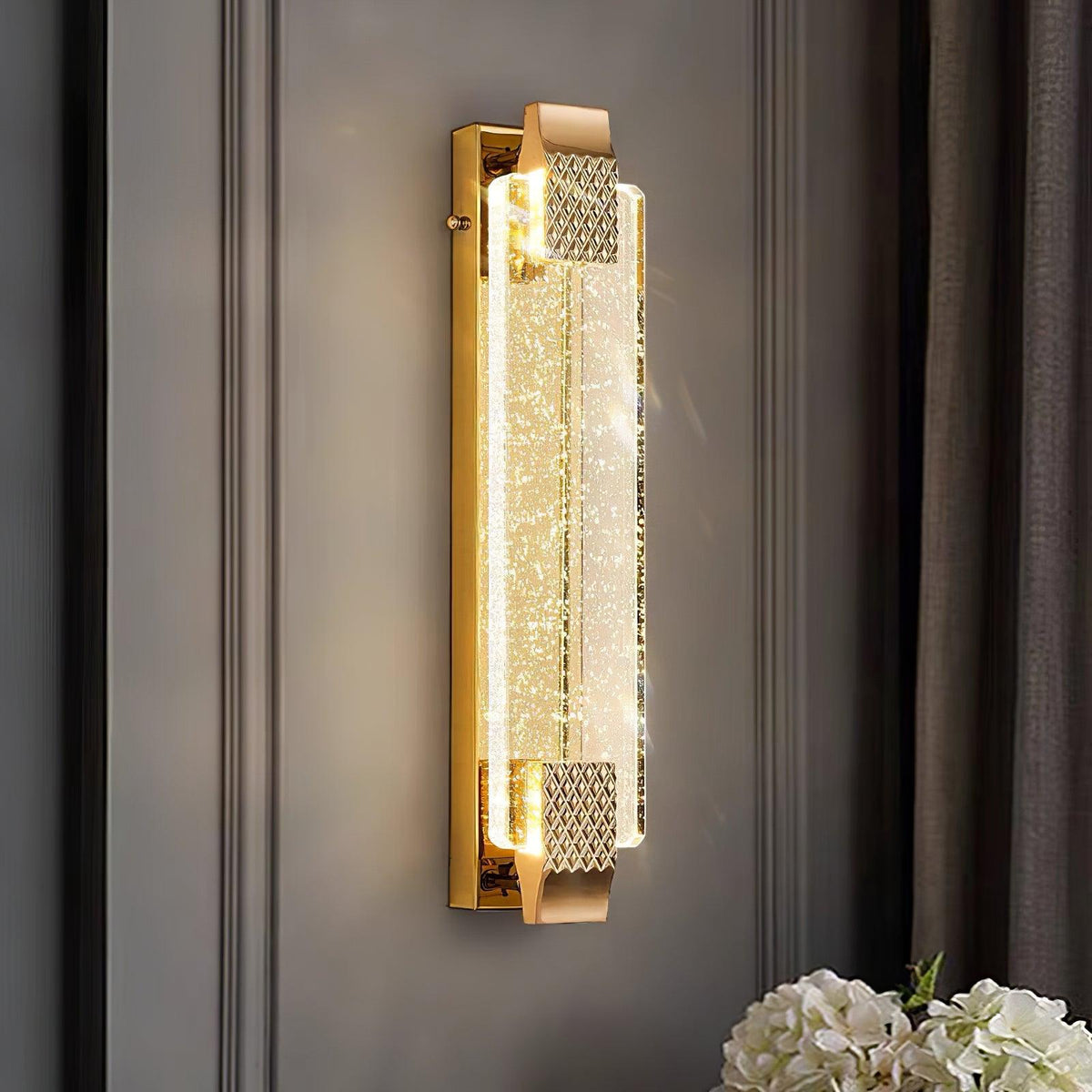 Lit bubble-crystal wall light from Clérachic on a grey panelled wall beside dark curtains, with white flowers below.