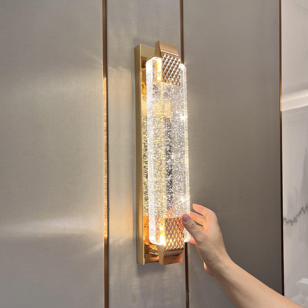 A hand rests beside the glowing bubble-crystal panel of this Clérachic wall light, mounted on a grey panelled wall.
