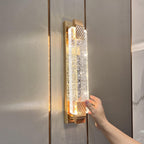 A hand rests beside the glowing bubble-crystal panel of this Clérachic wall light, mounted on a grey panelled wall.