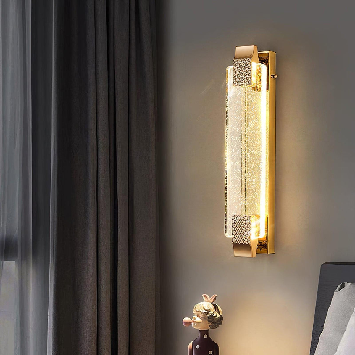 Warm-lit Clérachic crystal wall light on a grey bedroom wall next to heavy curtains, with a small figurine below.