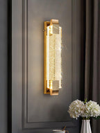 Clérachic crystal wall light glowing on a dark grey panelled wall beside a curtain, with white hydrangeas on a table below.
