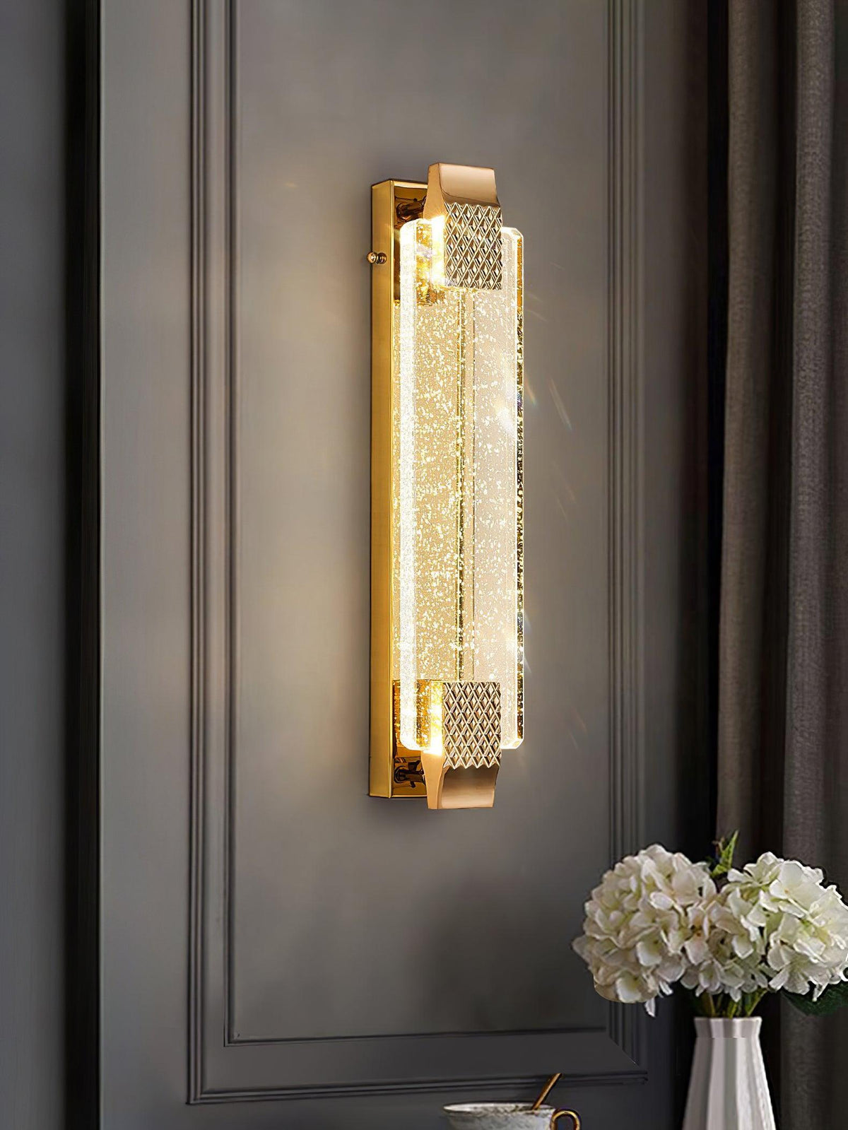 Clérachic crystal wall light glowing on a dark grey panelled wall beside a curtain, with white hydrangeas on a table below.