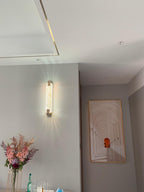 Clérachic wall light glowing on a pale wall in a hallway, with a framed arch print and pink flowers nearby.