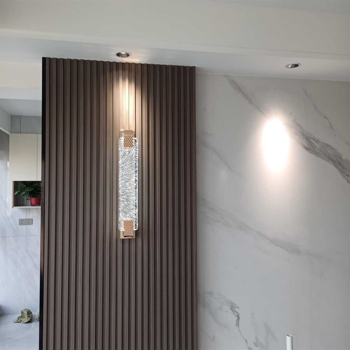 Clérachic crystal wall light mounted on a dark fluted panel next to a white marble wall and ceiling downlights.