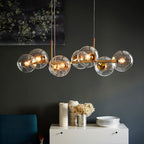 Gold Clérachic bubble glass linear pendant light with clear globes, hanging above a console styled with vases and floral art.